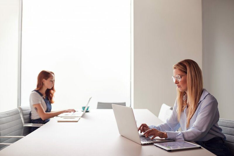 Two Casually Dressed Young Businesswomen Working On Laptops In Modern Meeting Room